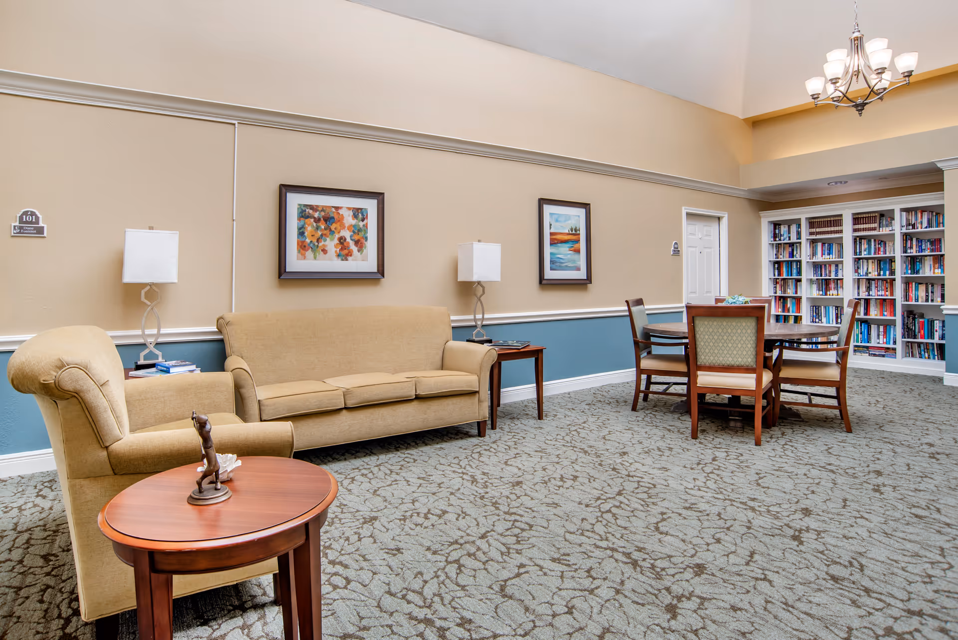 A cozy senior living facility common area with a beige sofa and armchair, two table lamps, framed artwork on the walls, a round wooden table with four chairs, and a large bookshelf filled with books. The room has a patterned carpet and soft lighting from a chandelier.
