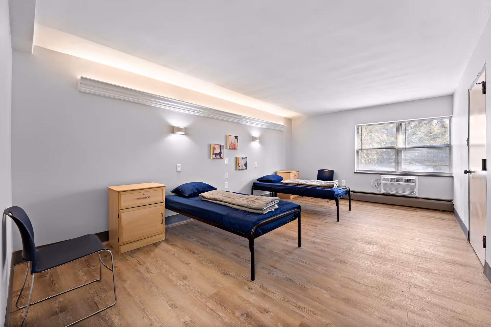 A simple, clean bedroom with two single beds each with navy blue bedding and a folded beige blanket on top. Each bed has a light wood nightstand beside it. There are two dark chairs, one near each nightstand. The room has light wood flooring, white walls, and a large window with blinds letting in natural light. Above the beds, there are three small abstract paintings and two wall-mounted lights with a decorative molding and indirect lighting near the ceiling.