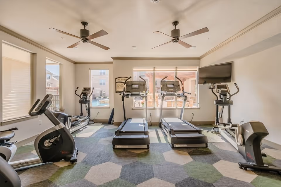 A bright fitness room with treadmills, ellipticals and stationary bikes facing windows and ceiling fans overhead.