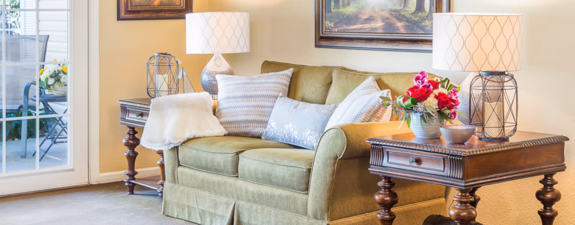 A cozy living room with a green upholstered sofa adorned with multiple decorative pillows and a white throw blanket. On either side of the sofa are wooden side tables with ornate legs, each holding a lamp with a patterned white lampshade. One side table also has a decorative metal candle holder, while the other has a vase with a colorful bouquet of flowers and a small bowl. A framed painting hangs on the beige wall above the sofa, and a glass door to the left reveals an outdoor patio area with chairs and a flower arrangement.