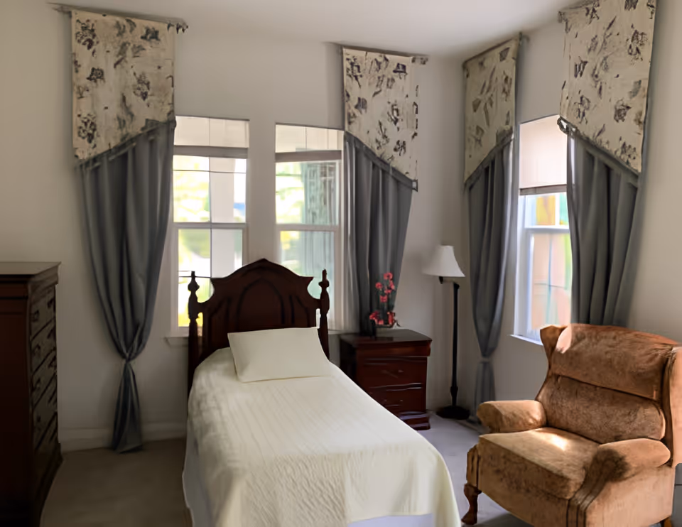 A cozy bedroom with a single bed covered in a white bedspread and a pillow. The bed has a dark wooden headboard and is positioned between two windows with gray curtains and patterned valances. To the right of the bed is a wooden nightstand with a small vase of pink flowers and a floor lamp behind it. A brown upholstered armchair is placed near the window on the right side of the room. There is also a wooden dresser on the left side of the room.