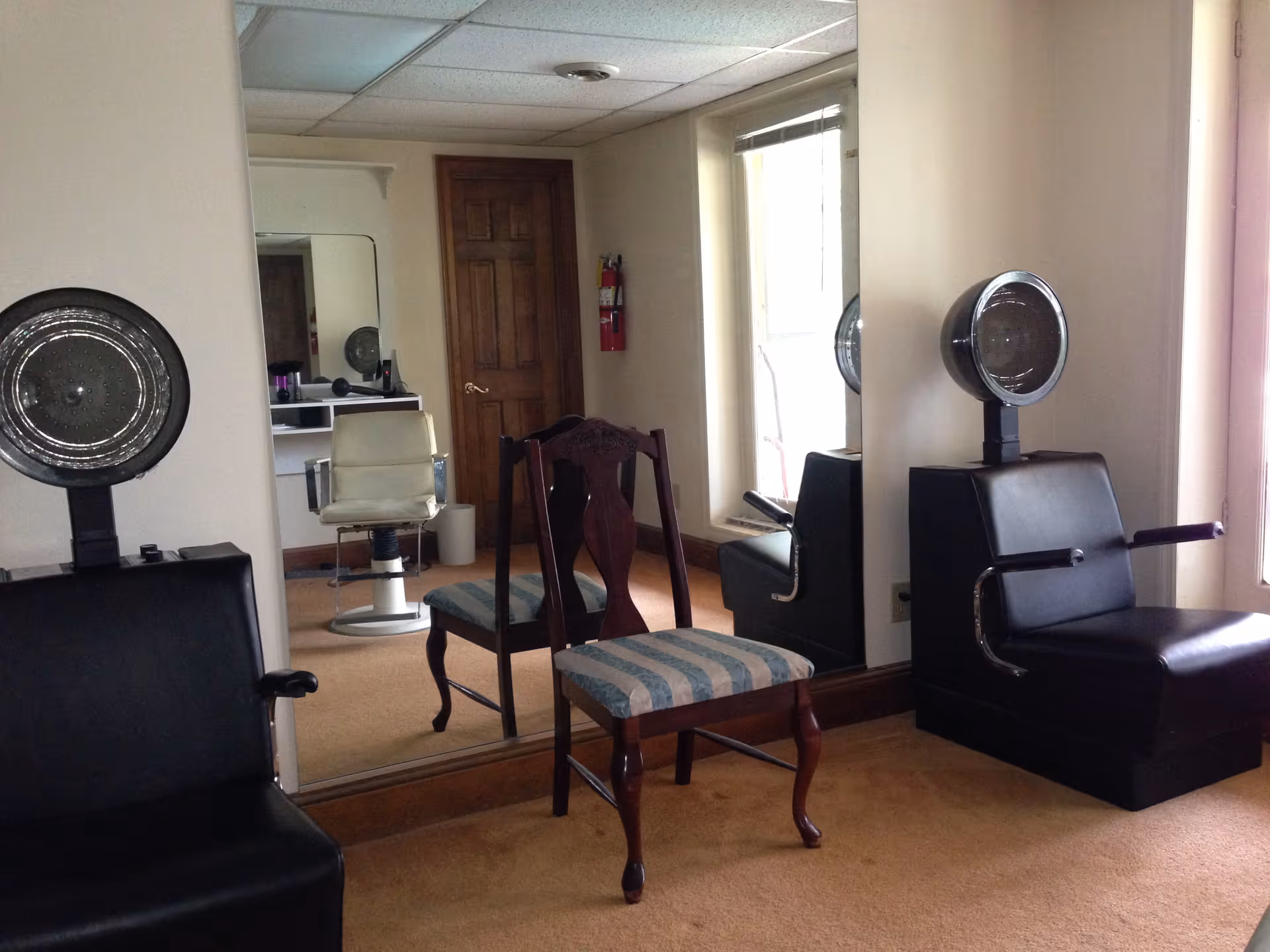 Interior view of a salon area with two black hair drying chairs facing a large mirror, a wooden chair with a striped cushion in front of the mirror, and a white salon chair visible in the reflection. There is a wooden door and a fire extinguisher on the wall in the background.