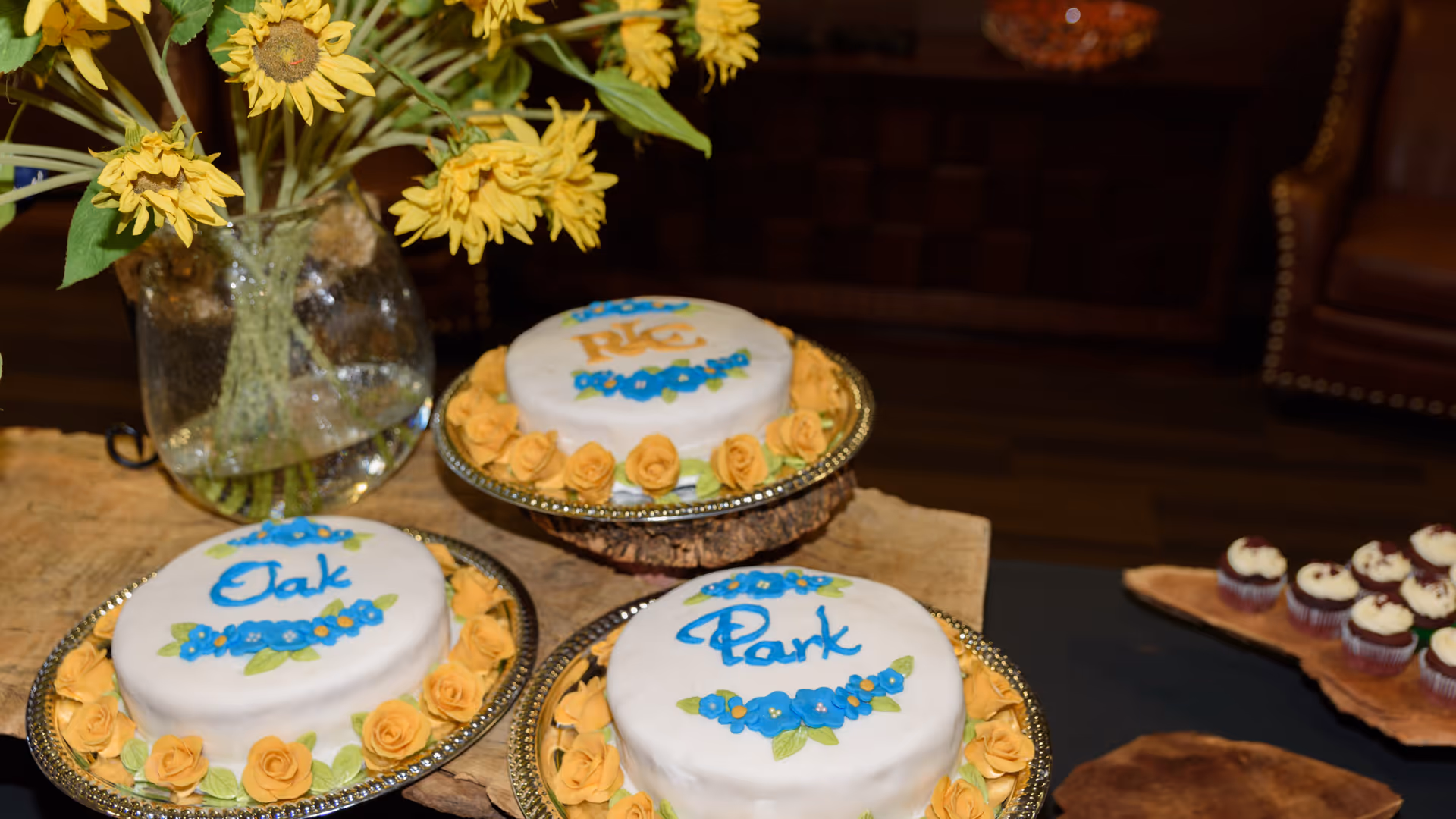 Three round white cakes decorated with yellow roses and blue flowers, each with the words 'Oak', 'Park', and 'RC' written in blue icing, displayed on silver trays on a wooden table. A vase with yellow sunflowers is placed behind the cakes, and a wooden tray with cupcakes is visible to the right.