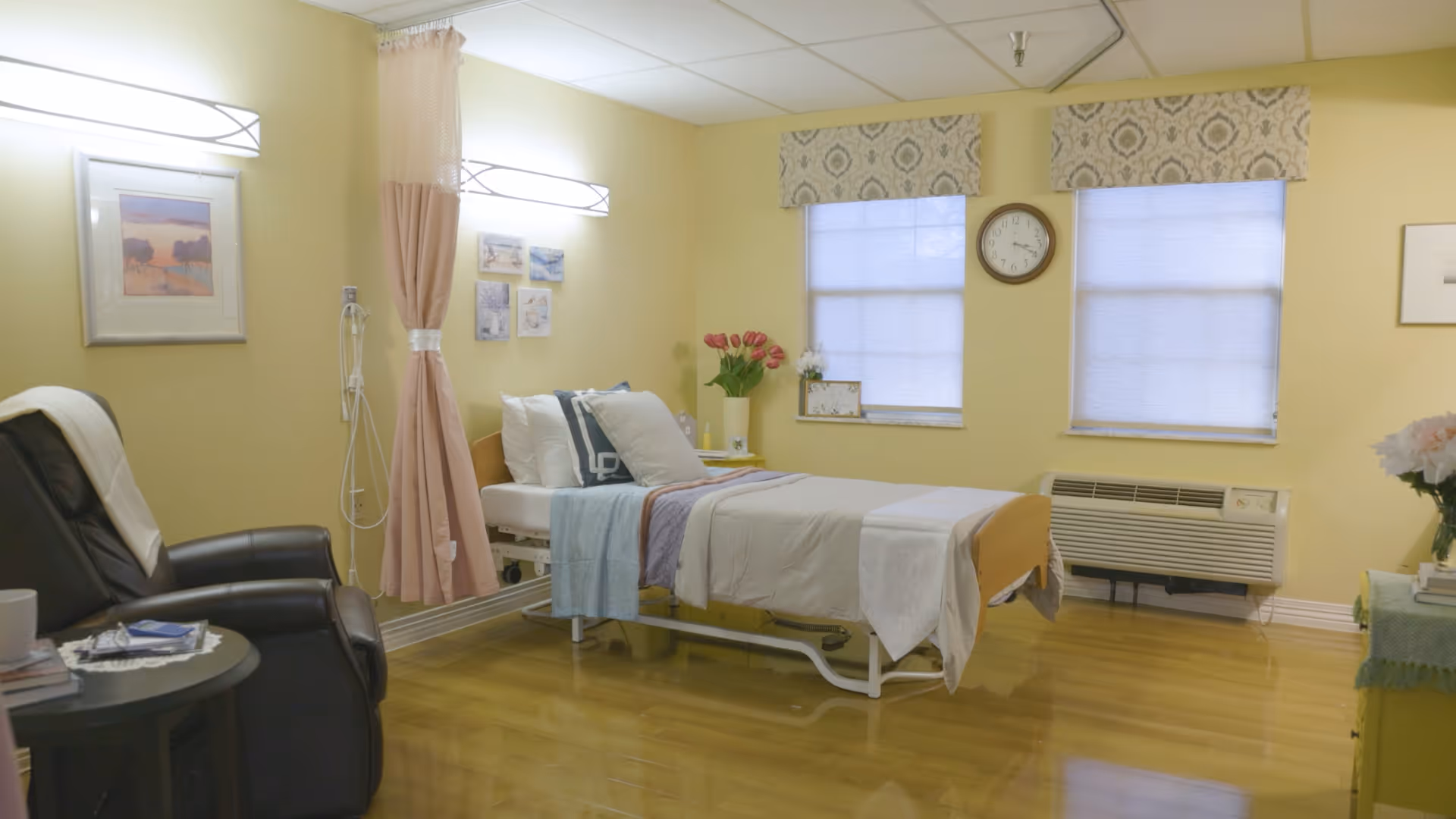 A neatly arranged bedroom in a healthcare facility with a single hospital bed made up with white and pastel bedding. There is a black recliner chair next to a small round table with a cup and some reading materials. The walls are painted light yellow and decorated with framed pictures. Two windows with patterned valances and blinds are visible, along with a wall clock and an air conditioning unit beneath the windows. A vase with pink flowers is placed on a small table near the bed.