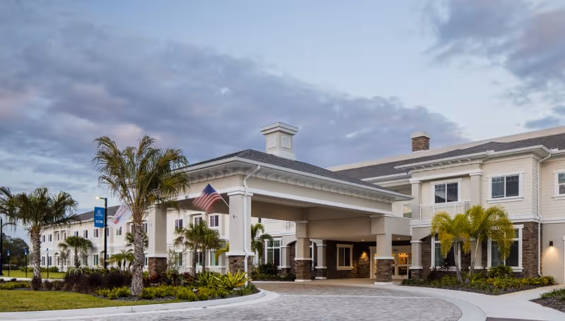 Exterior view of American House St. Petersburg senior living facility showing a large covered entrance with columns, palm trees, and landscaped greenery under a cloudy sky at dusk.