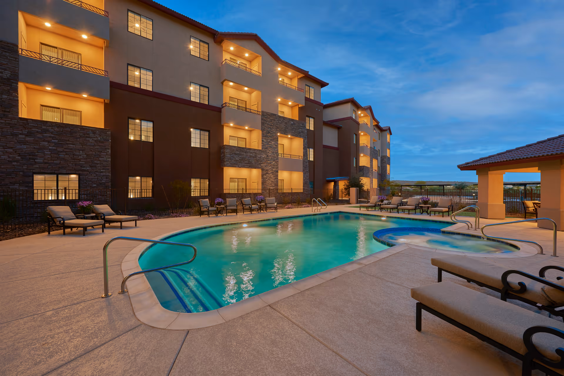Outdoor swimming pool area at dusk with lounge chairs and tables surrounding the pool. A multi-story residential building with balconies and warm lighting is visible in the background under a partly cloudy sky.