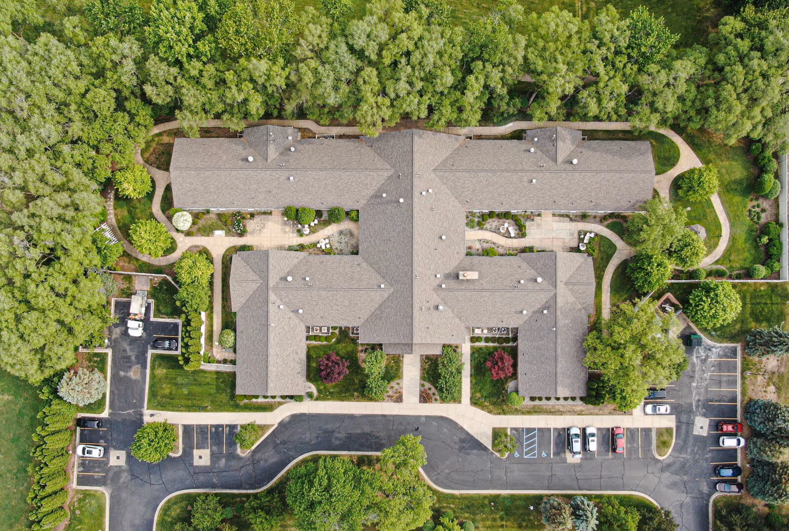 Aerial view of Arden Courts - ProMedica Memory Care Community in Sterling Heights showing a large building with a complex roof structure surrounded by trees, landscaped gardens, walking paths, and parking areas with several cars.
