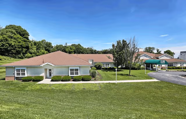 Exterior view of a single-story senior living facility building with a brown roof, white siding, and several windows. The building is surrounded by a well-maintained lawn, bushes, and trees under a clear blue sky. A paved driveway leads to a covered entrance on the right side.