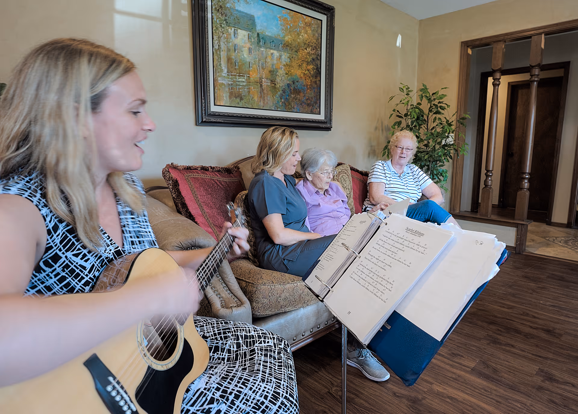 A woman playing an acoustic guitar and singing from sheet music on a stand, while three elderly women sit on a couch in a cozy living room with a painting on the wall and a potted plant nearby.