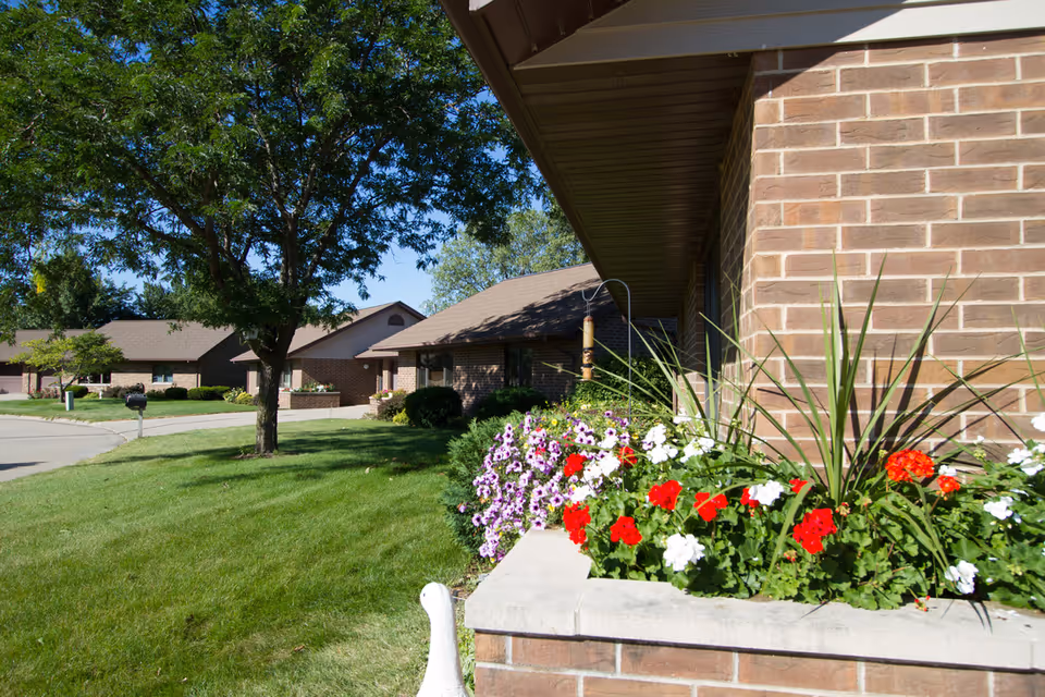 Brick single-story residential units with a flower planter in the foreground, lawn, and trees.