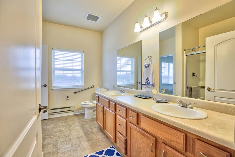 Bright bathroom with a double sink vanity featuring wooden cabinets and a large mirror above. There is a window with white blinds, a toilet with a grab bar beside it, and a shower with a glass door. The floor has beige tiles and a blue patterned rug is placed in front of the sinks.