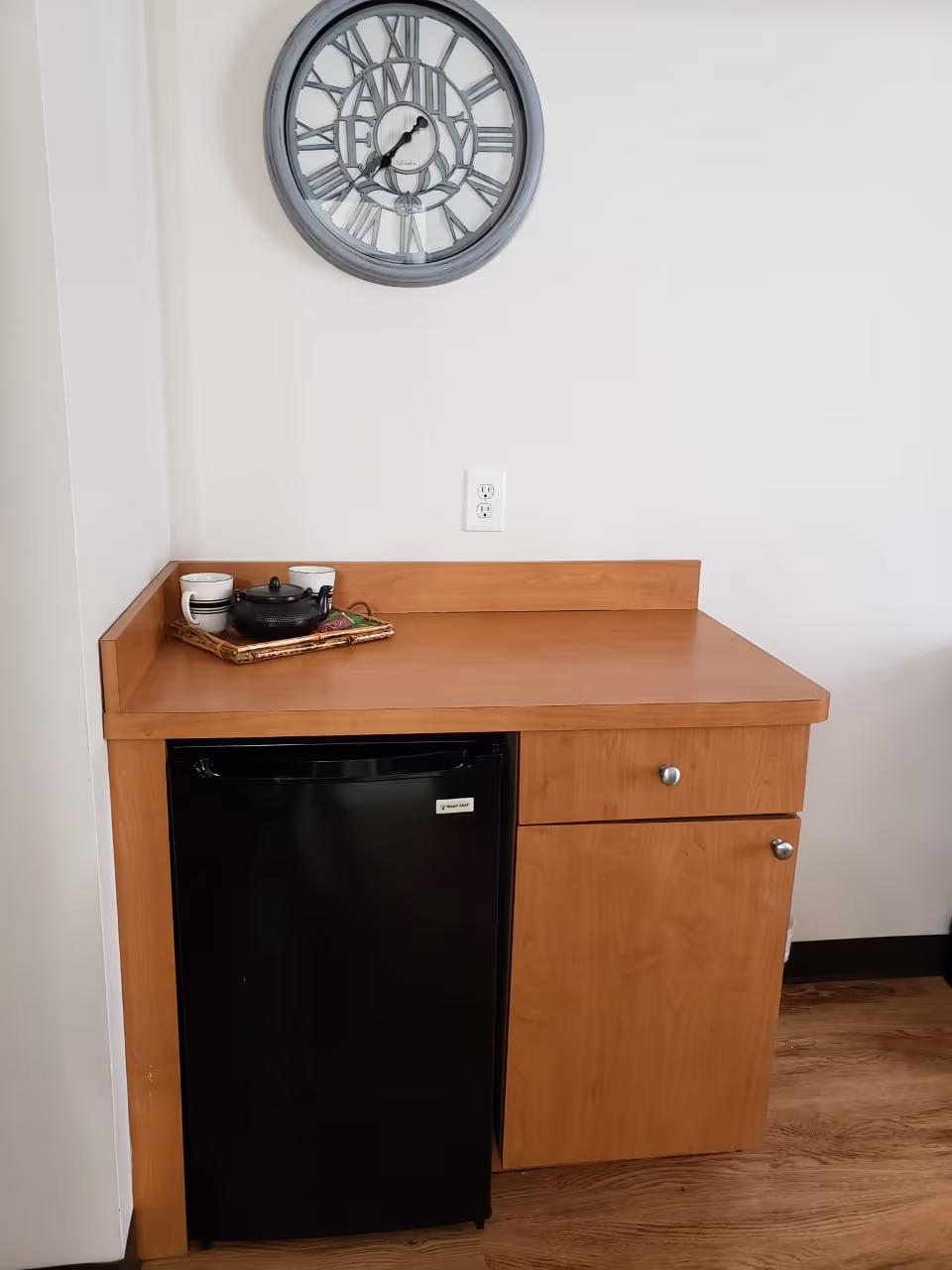 A small wooden kitchenette area with a black mini refrigerator, a drawer, and a cabinet. On the countertop, there is a wicker tray holding a black teapot and two white cups. Above the kitchenette, a round wall clock with the word FAMILY in the center is mounted on a white wall.