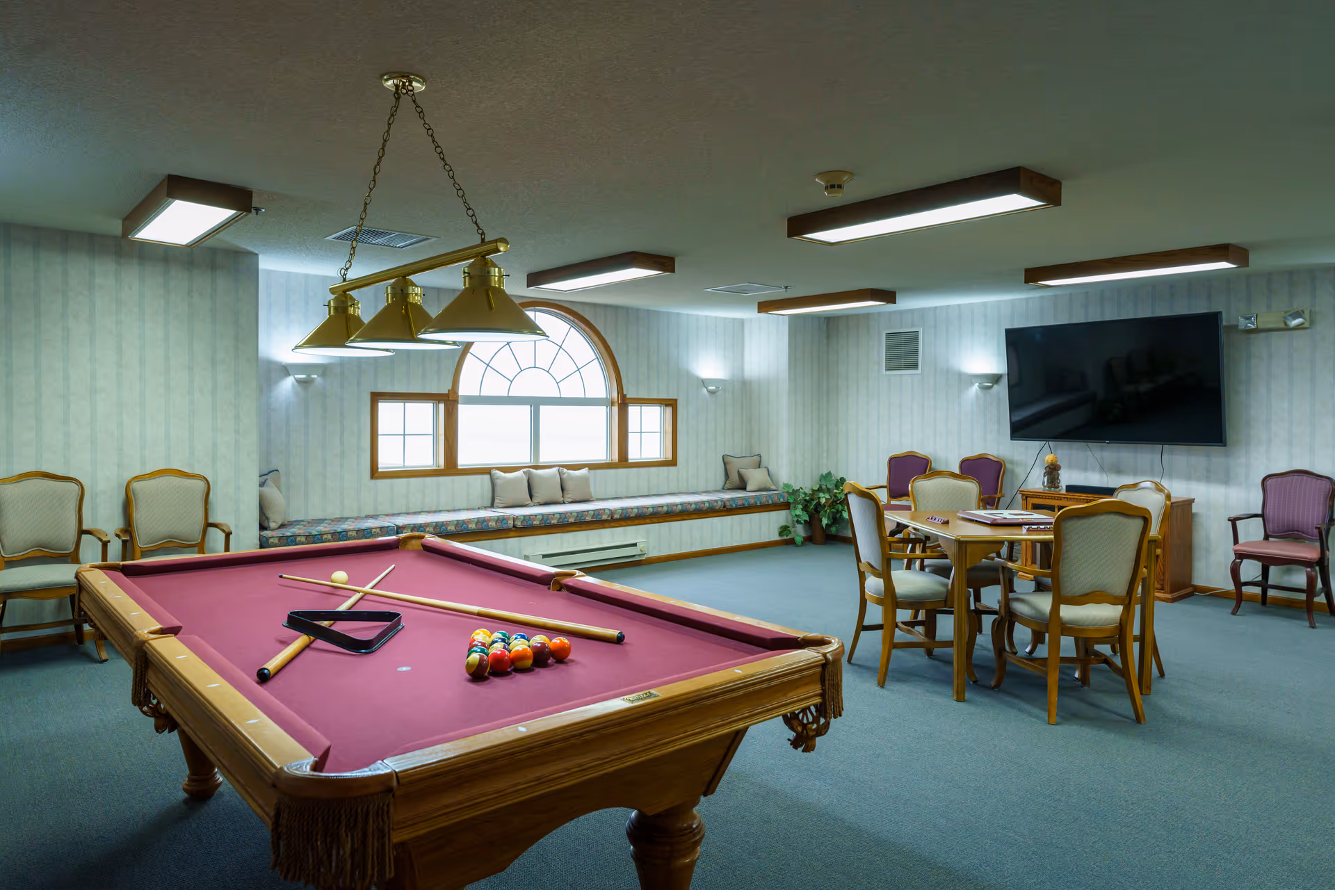 A recreational room with a pool table featuring a red felt surface and pool cues arranged on it. The room has a large arched window with a cushioned bench underneath, several chairs, a round table with chairs, a flat-screen TV mounted on the wall, and soft lighting fixtures on the ceiling and walls.