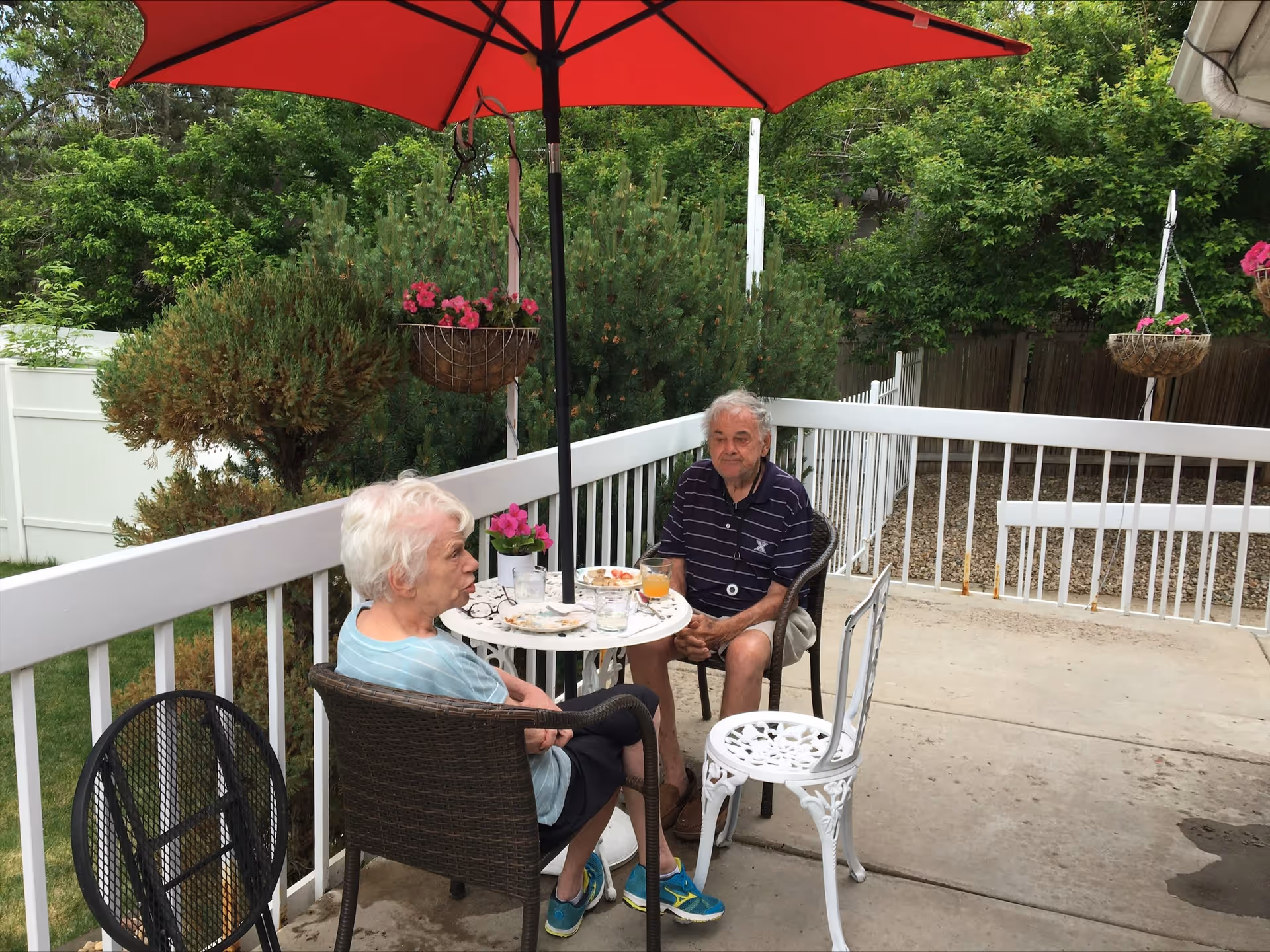 Two elderly people sitting at a small round table with drinks and snacks on a patio. They are seated under a large red umbrella, surrounded by greenery and hanging flower baskets. The patio has a white railing and concrete floor.