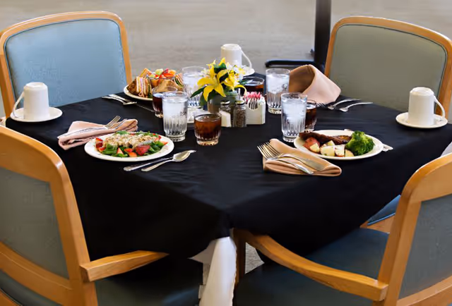 Round dining table set for four with plated meals, glasses, napkins, and a small floral centerpiece in a dining area.