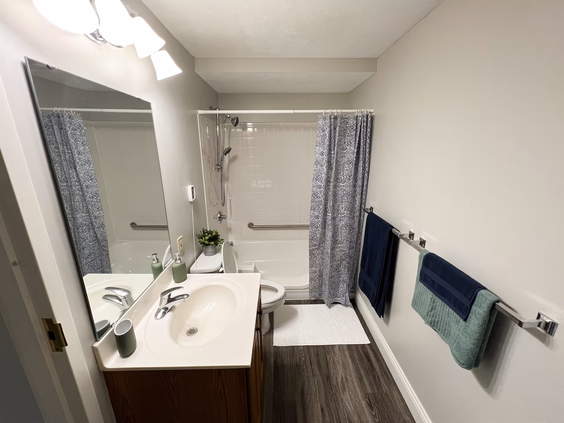 A clean and modern bathroom featuring a white sink with a silver faucet, a large mirror above the sink, a toilet with the lid open, and a bathtub with a showerhead and a patterned shower curtain. The bathroom has light-colored walls, dark wood flooring, two towel racks with blue and green towels, and a small potted plant on the toilet tank.
