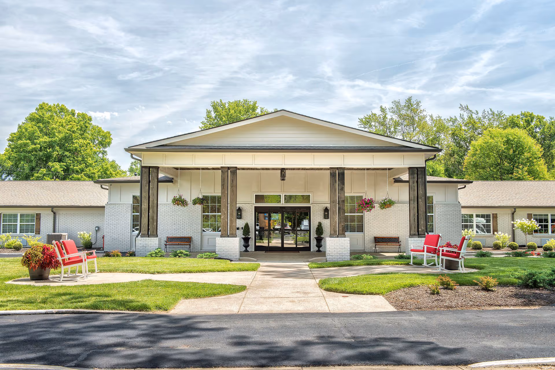 Front exterior view of a single-story senior living facility building with a covered entrance supported by wooden pillars. There are hanging flower baskets, benches, and red cushioned chairs on the landscaped lawn with green grass and shrubs. Trees and a partly cloudy sky are visible in the background.