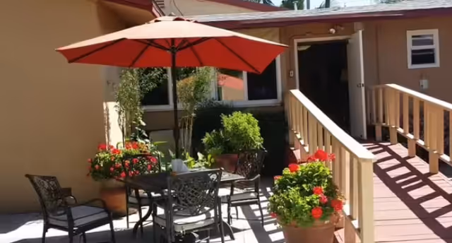 Outdoor patio area with a round metal table and four chairs under a large red umbrella. The patio is surrounded by potted plants with red flowers and greenery. A wooden ramp leads to a door of a beige building in the background.