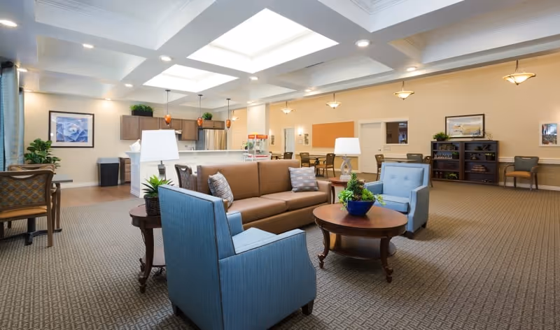 Spacious communal lounge with a brown sofa and blue armchairs around a coffee table and an open kitchenette in the background.