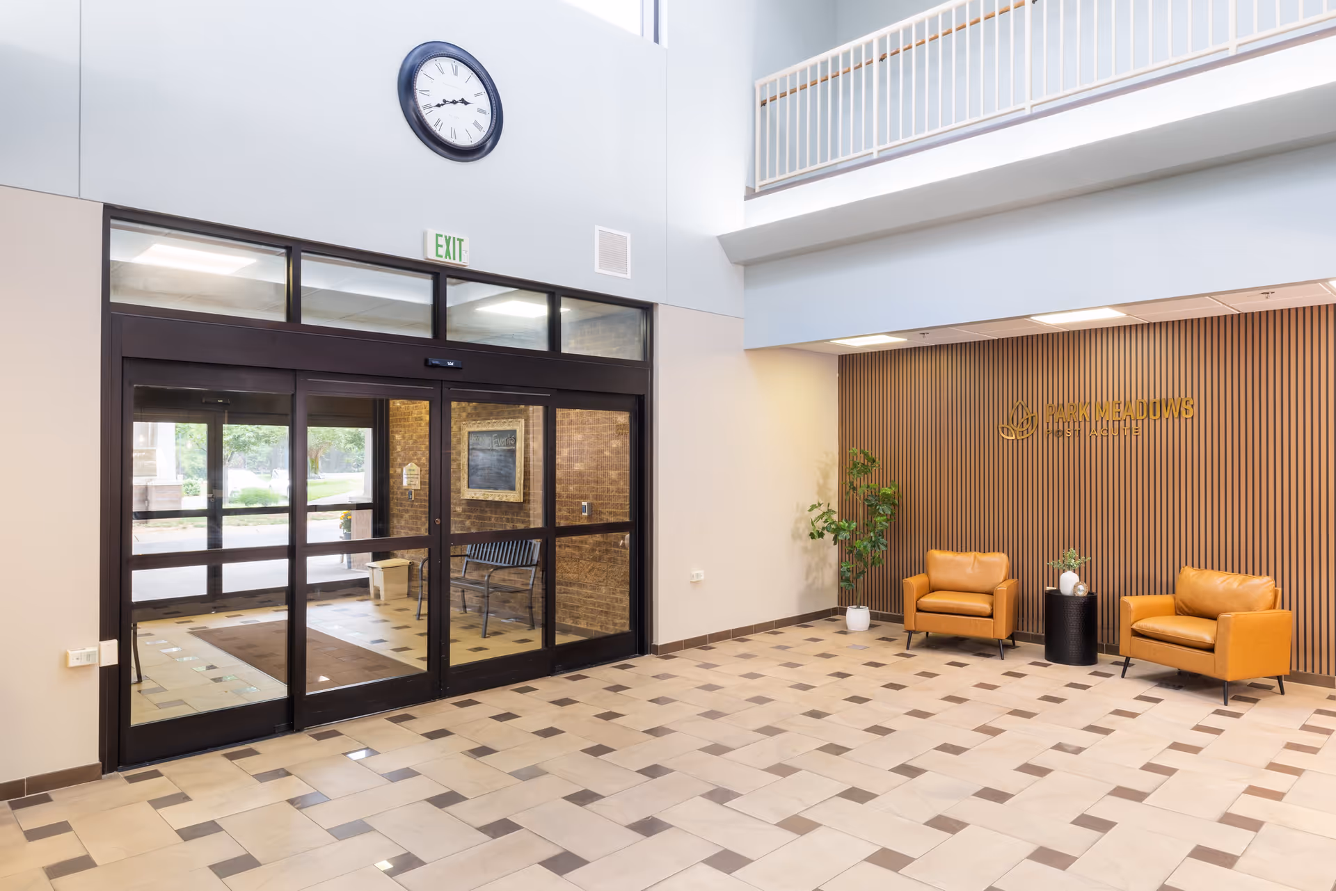 Spacious lobby area with large glass entrance doors, a clock on the wall above the doors, two tan leather chairs with a small black table between them, a potted plant, and a wooden slat wall with the Park Meadows Post Acute logo.