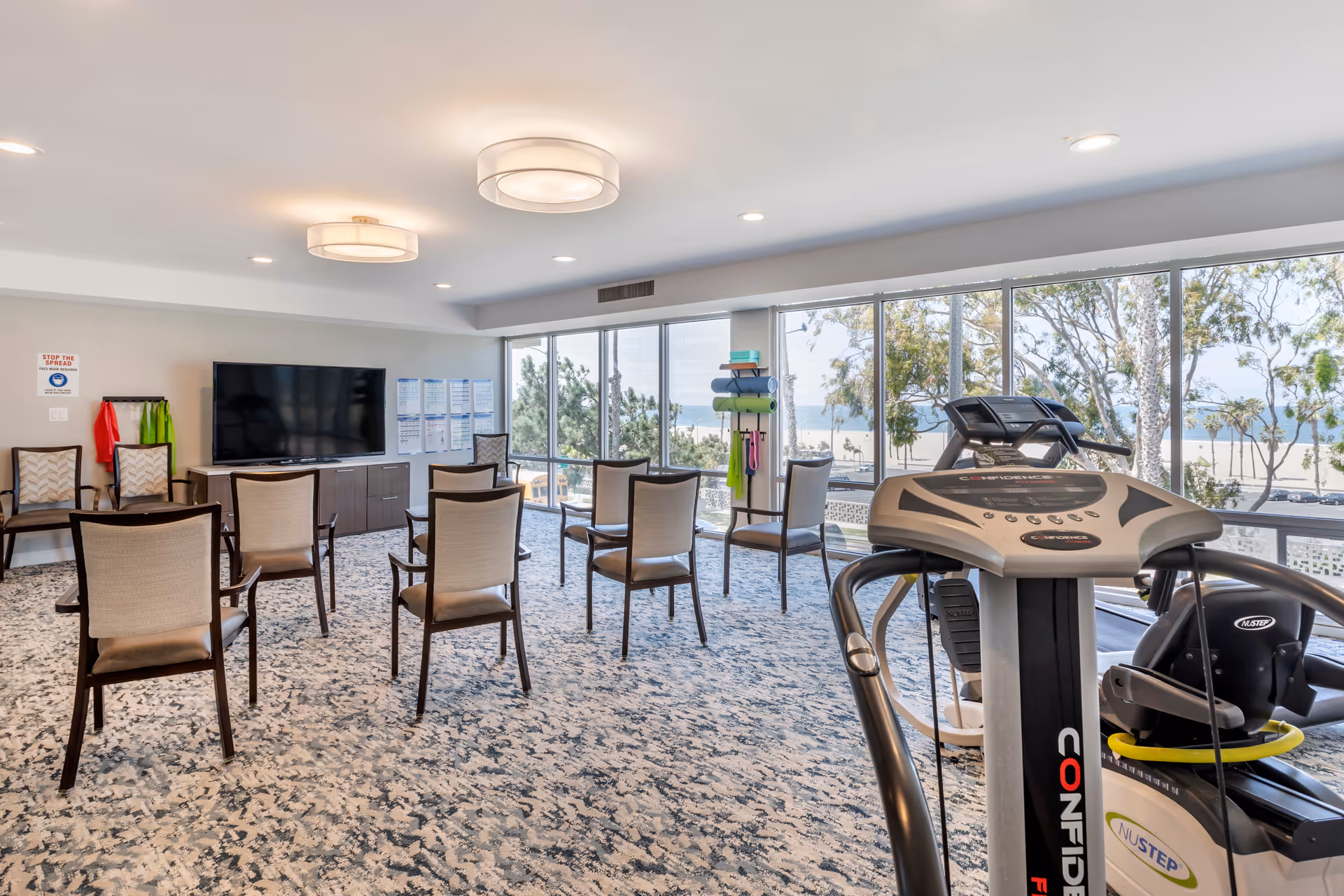 Bright activity room with rows of chairs facing a TV and exercise equipment by floor-to-ceiling windows overlooking the beach.