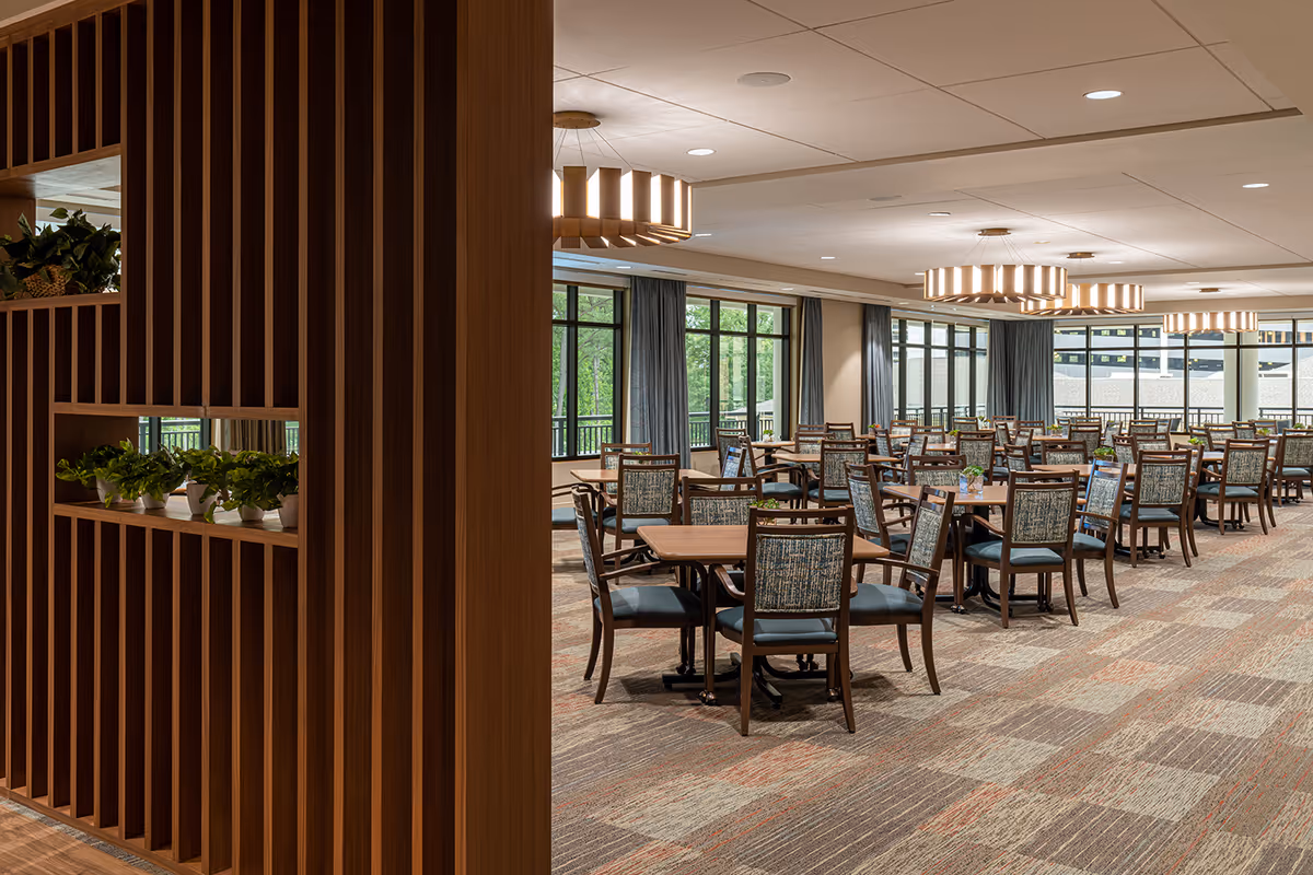 A spacious dining room with multiple wooden tables and chairs arranged neatly. The room features large windows with blue curtains allowing natural light to brighten the space. Modern circular chandeliers hang from the ceiling, and a wooden partition with small potted plants is visible on the left side.