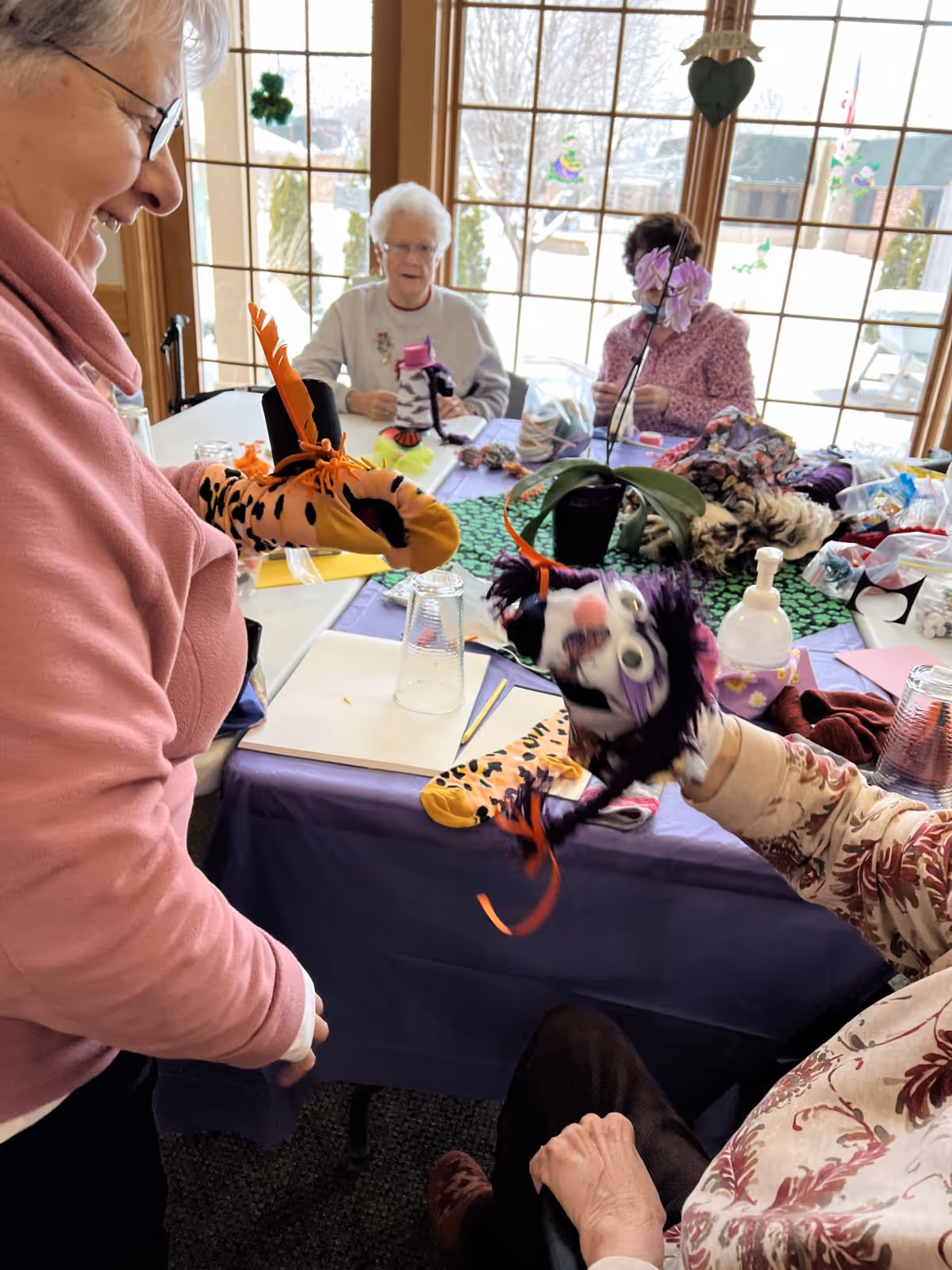 Two elderly women sitting at a table engaged in a craft activity with colorful sock puppets, while another woman stands nearby smiling and interacting with them. The room has large windows with a snowy outdoor view and various craft supplies on the table.