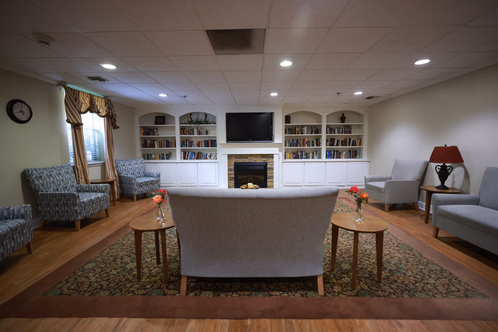 A cozy living room area in a senior living facility with a central fireplace and a mounted TV above it. The room features built-in white bookshelves filled with books on either side of the fireplace. There are multiple upholstered chairs and a sofa arranged around a patterned area rug, with small wooden tables holding vases with flowers. A window with curtains and a wall clock are visible on the left side, and a table lamp with a red shade is on the right.