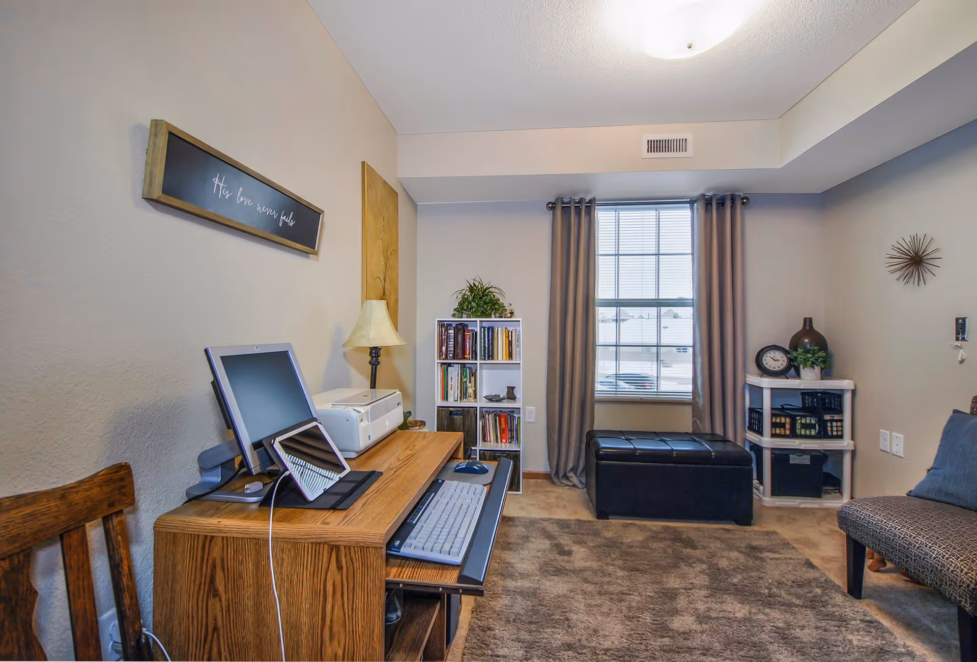 A cozy room with a wooden desk holding a computer monitor, keyboard, mouse, and printer. A wooden chair is positioned next to the desk. Behind the desk is a small bookshelf filled with books and a plant on top. A window with beige curtains lets in natural light. In front of the window is a black cushioned storage bench. To the right, there is a small white shelving unit with decorative items including a clock and a plant. A cushioned chair with a pillow is partially visible on the right side. The walls are light beige, and a framed sign with cursive writing hangs above the desk.