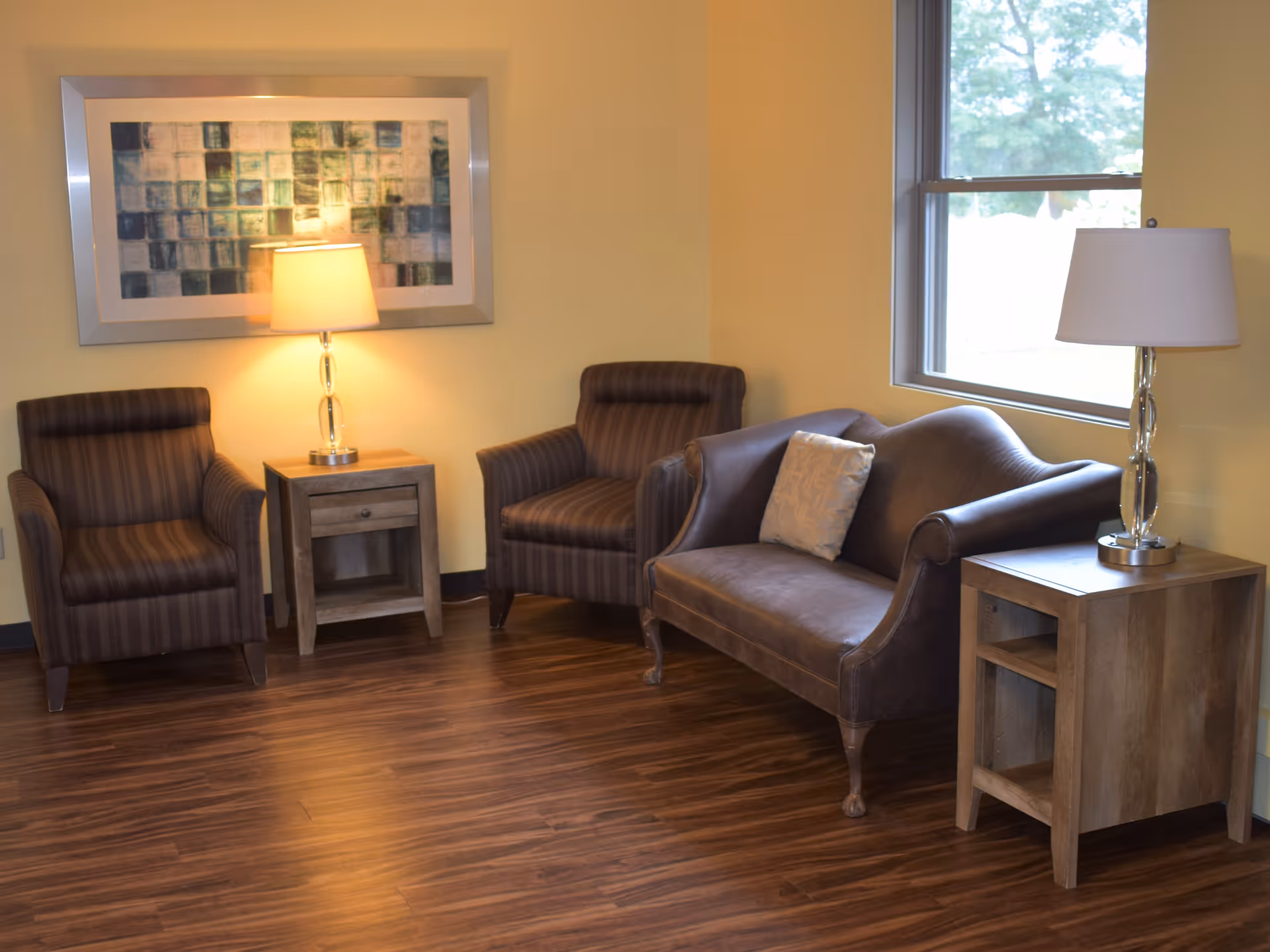 A cozy sitting area with two striped armchairs and a brown leather loveseat with a decorative pillow. There are two wooden side tables, each with a lamp, and a large window letting in natural light. A framed abstract artwork hangs on the yellow wall above one of the lamps.