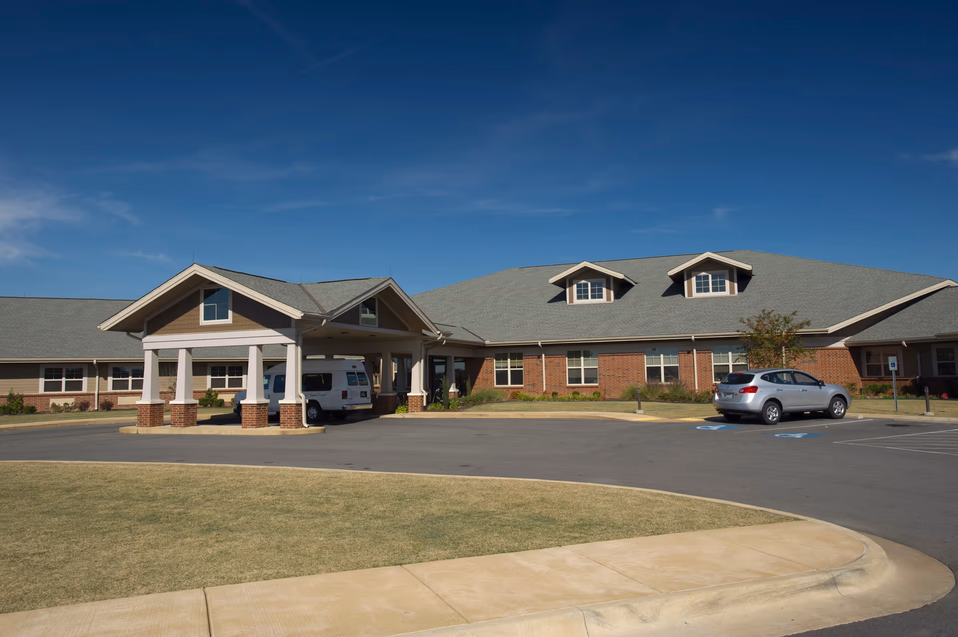 Exterior view of a single-story brick building with a covered entrance and a driveway. There are a few parked vehicles, including a white van under the entrance canopy and a silver car in a parking space. The sky is clear and blue.