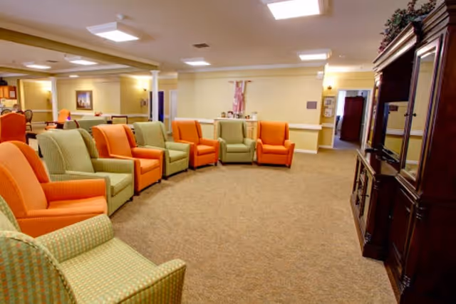 A spacious senior living facility common area with a semi-circle arrangement of orange and green armchairs facing a large wooden entertainment center with a television. The room has beige walls, carpeted floors, and bright ceiling lights, creating a warm and inviting atmosphere.