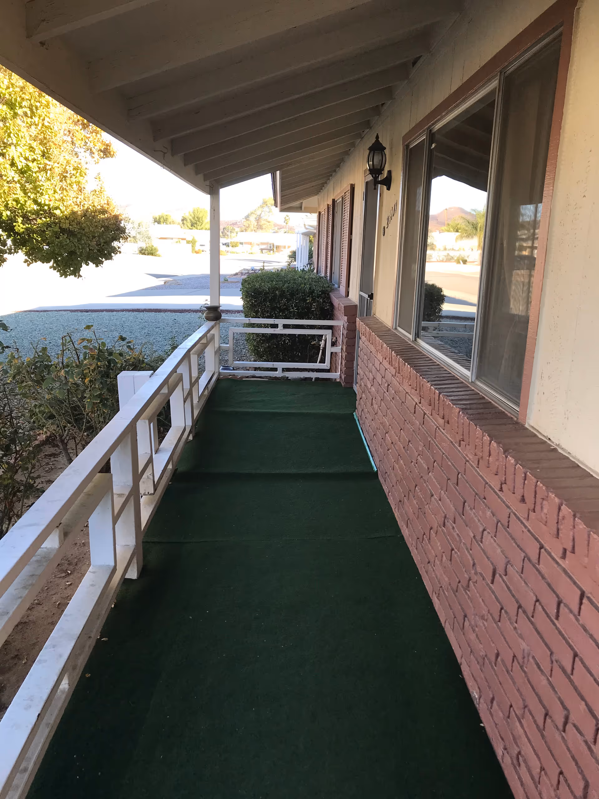 Covered front porch with green outdoor carpet, white railing, and a brick exterior wall under a slanted roof.