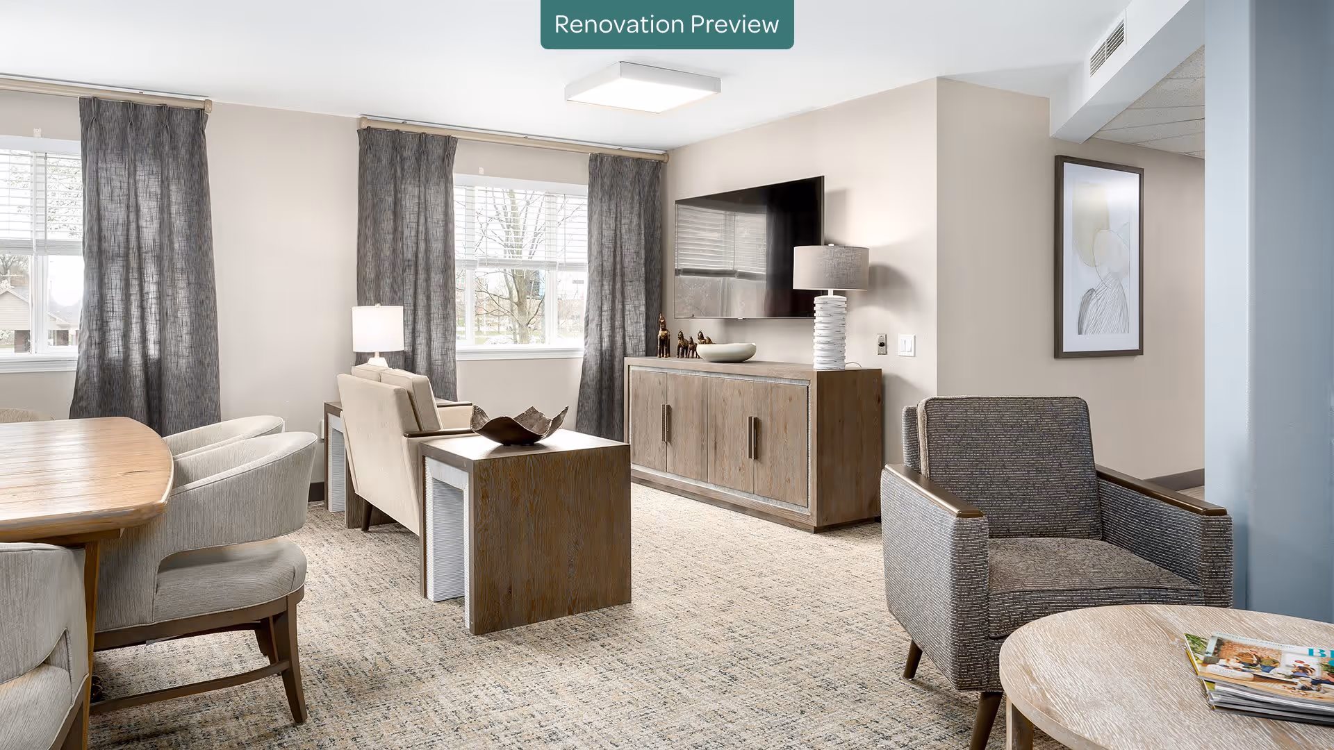 A bright and modern living room with large windows covered by gray curtains, a wooden dining table with upholstered chairs, two armchairs, a wooden side table, a wooden media console with a flat-screen TV mounted above it, and a table lamp. The room has light-colored walls and carpeted flooring.