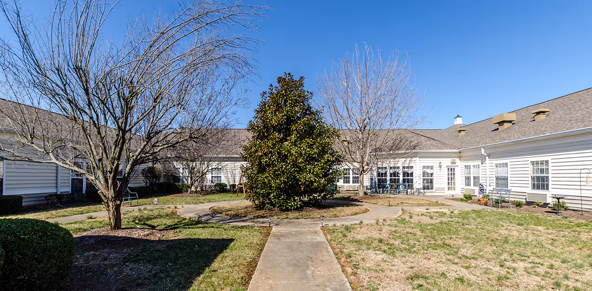 Outdoor courtyard area of The Bungalows at Bowling Green featuring a circular walkway around a large green tree, surrounded by leafless trees and white building walls with multiple windows and benches.