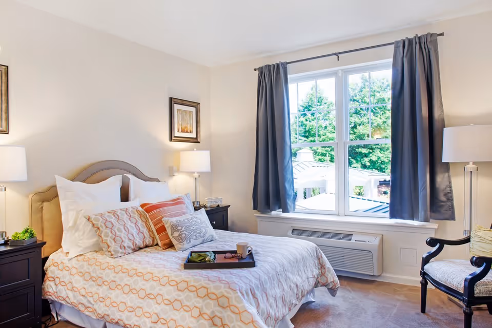 A bright and cozy bedroom with a neatly made bed featuring patterned bedding and multiple pillows. There are two bedside tables with lamps on each side of the bed. A large window with dark curtains lets in natural light and offers a view of greenery outside. A cushioned armchair is positioned near the window.