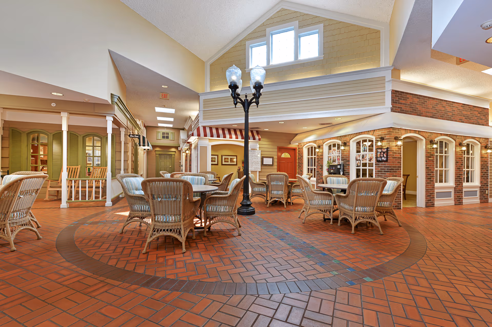 Bright indoor common area styled like a small village square with wicker tables and chairs, brick flooring, a central lamppost, and storefront-style facades.