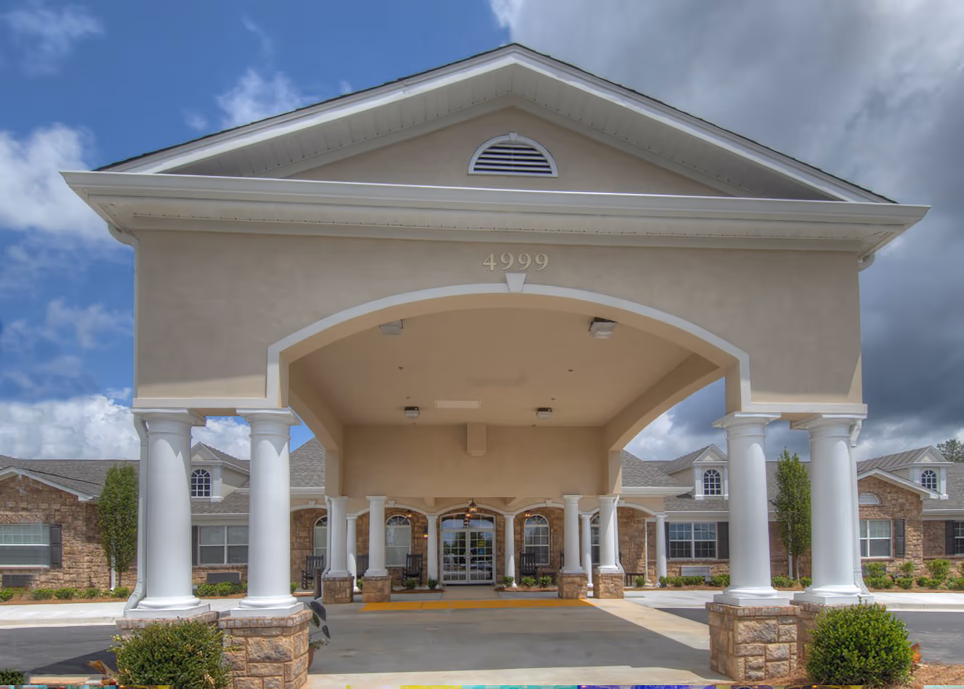 Front entrance of Benton House of Douglasville featuring a covered driveway supported by white columns with stone bases, beige exterior walls, and large windows under a partly cloudy sky.