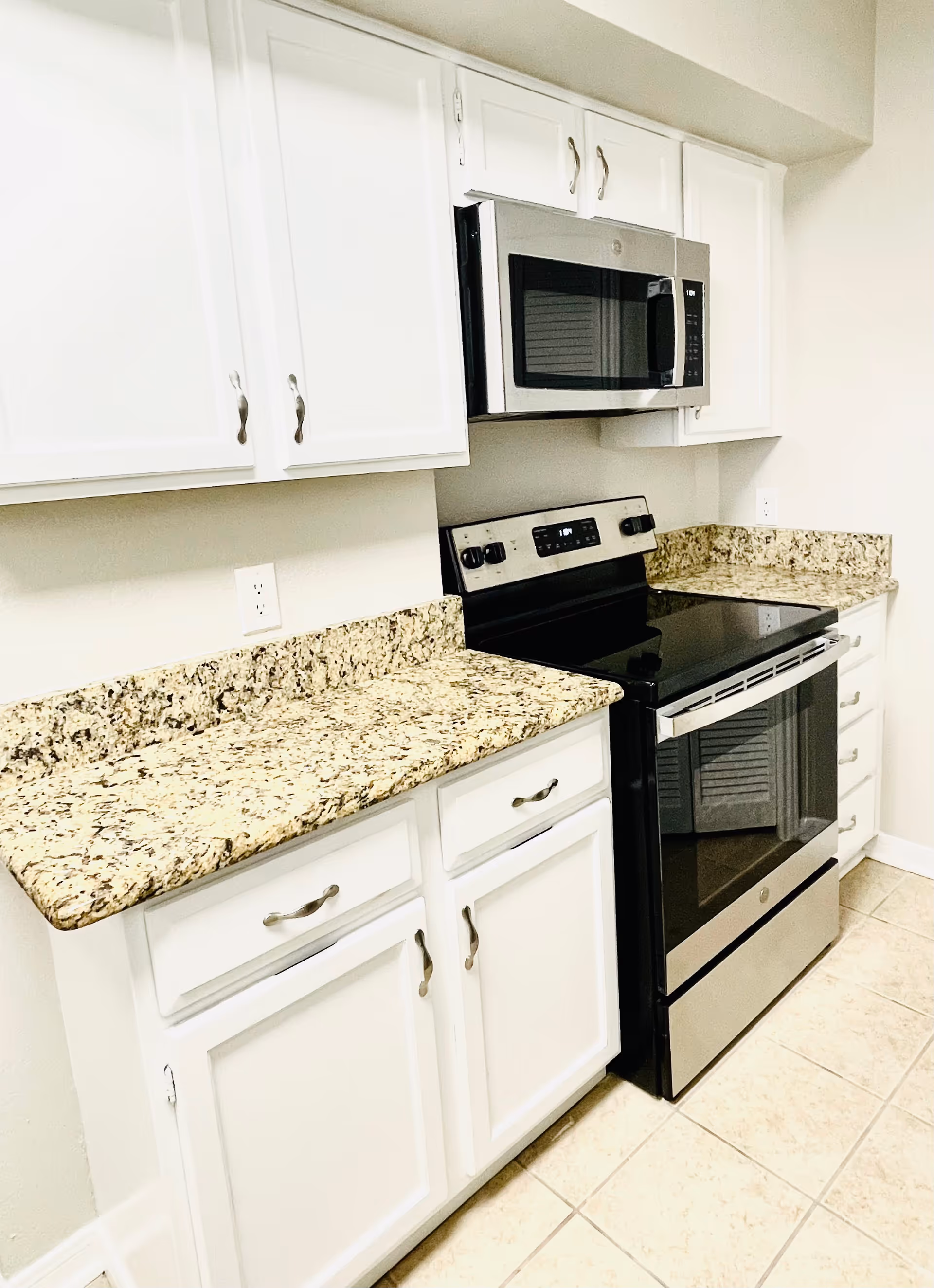 A modern kitchen area featuring white cabinets with silver handles, granite countertops, a stainless steel microwave mounted above a stainless steel electric stove, and beige tiled flooring.
