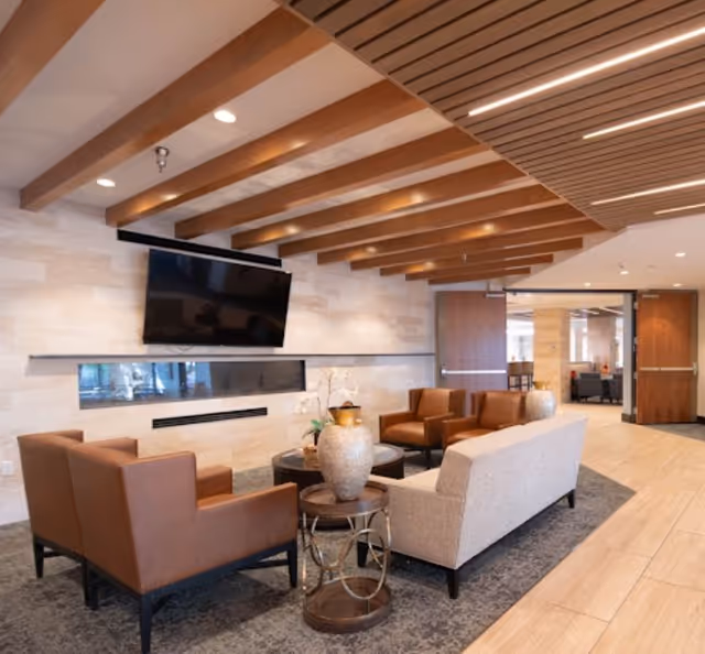 A modern and cozy living room area with a beige sofa, two brown armchairs, a round side table with a decorative vase, a wall-mounted flat-screen TV above a sleek fireplace, wooden ceiling beams, and light-colored tiled flooring.