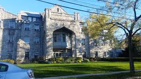 Stone building facade with arched entrance, well-kept lawn, trees, and a parked car.