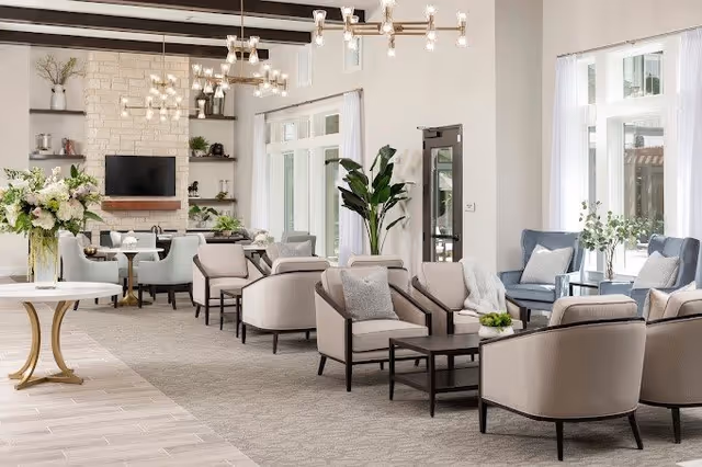 A spacious and well-lit living room area with multiple beige armchairs arranged around dark coffee tables. The room features large windows with white curtains, a stone fireplace with a mounted TV above it, modern chandeliers, and decorative plants and flowers.