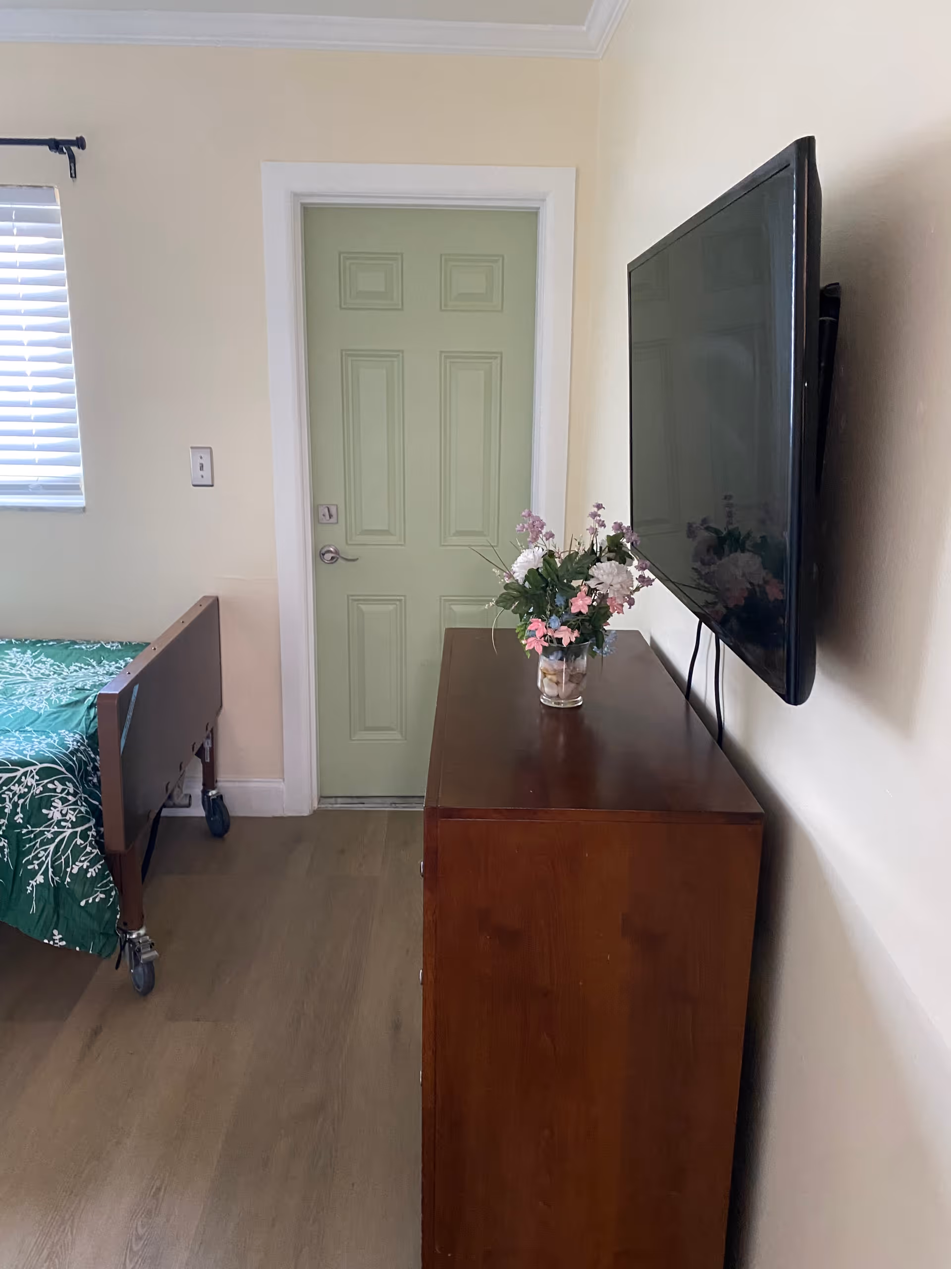 A small room with a hospital-style bed with a green floral bedspread, a wooden dresser with a vase of flowers on top, a wall-mounted flat screen TV, a light green door, and a window with closed blinds.