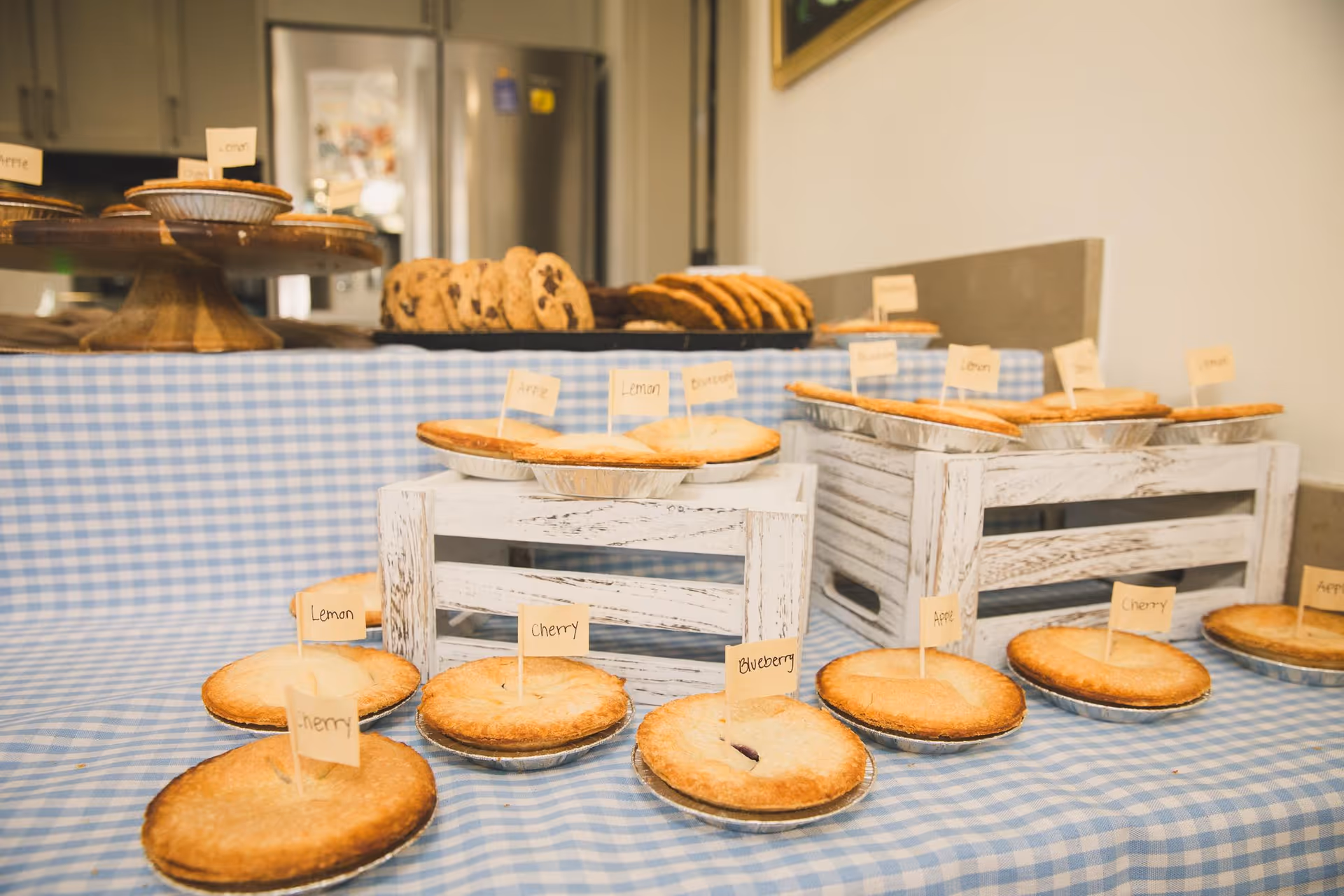 A display of various pies including lemon, cherry, blueberry, and apple, each labeled with small handwritten flags, arranged on white wooden crates and a blue and white checkered tablecloth. In the background, there are cookies and a refrigerator visible in a kitchen setting.