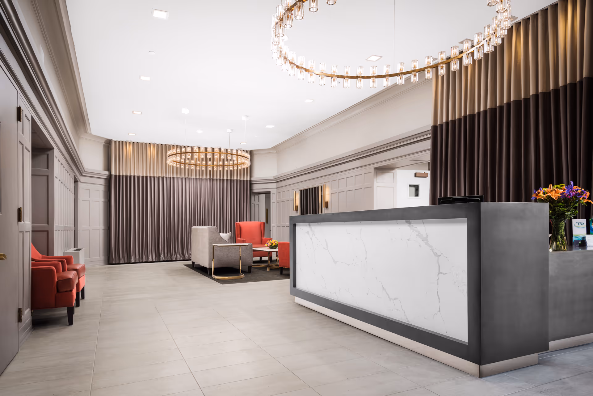 A modern and spacious reception area with a large marble-front desk on the right, a seating area with red and beige chairs in the background, elegant circular chandeliers hanging from the ceiling, and tall curtains covering the walls.