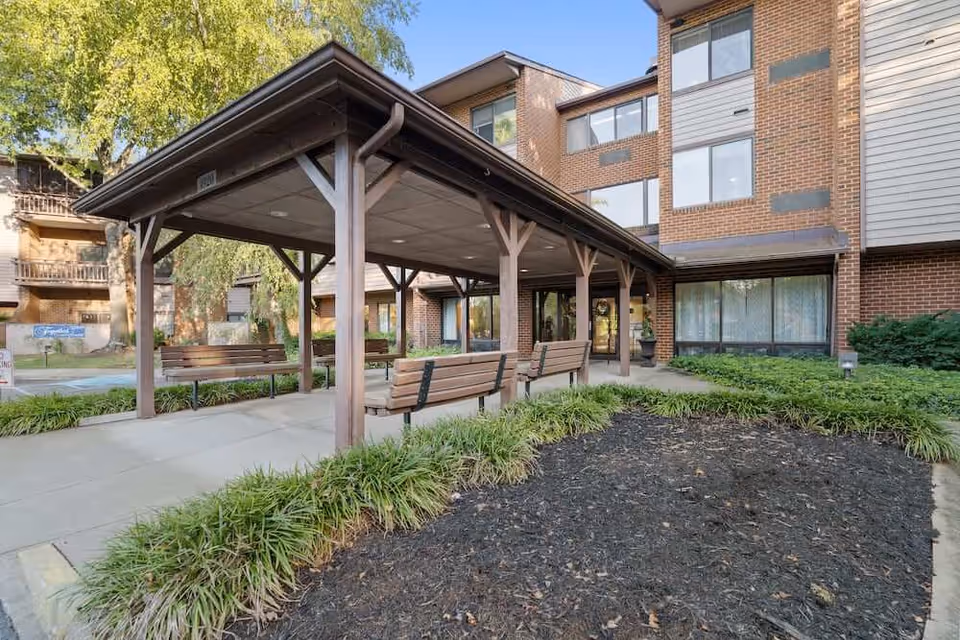 Covered outdoor seating area with wooden benches in front of a multi-story brick and siding building, surrounded by greenery and a landscaped garden bed.