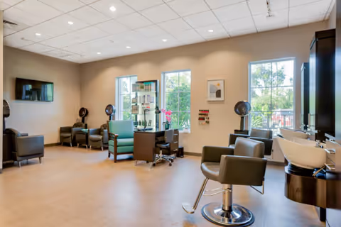Interior of a senior living facility salon area with multiple salon chairs, hair dryers, a TV mounted on the wall, and large windows letting in natural light.