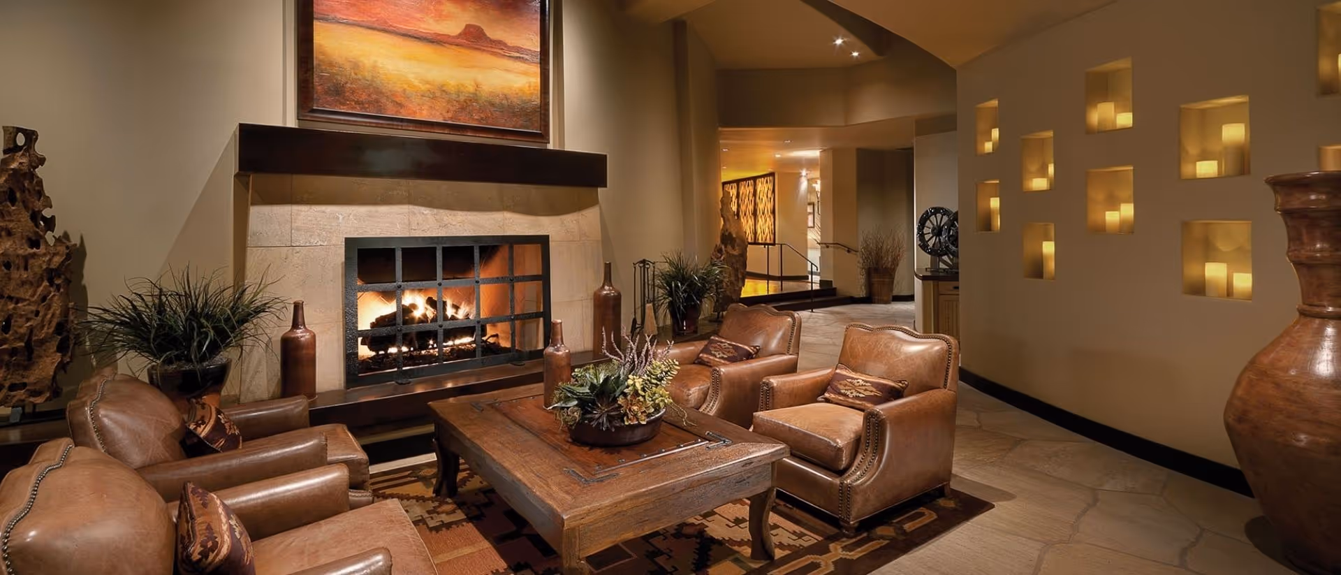 Cozy living room area with four brown leather armchairs arranged around a wooden coffee table. A fireplace with a fire burning is set in the stone wall, above which hangs a landscape painting. The room features warm lighting, decorative plants, large vases, and a wall with recessed candle niches.