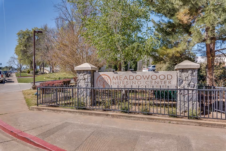 Stone entrance sign reading 'Meadowood Nursing Center' surrounded by trees, a metal railing, and parked cars.