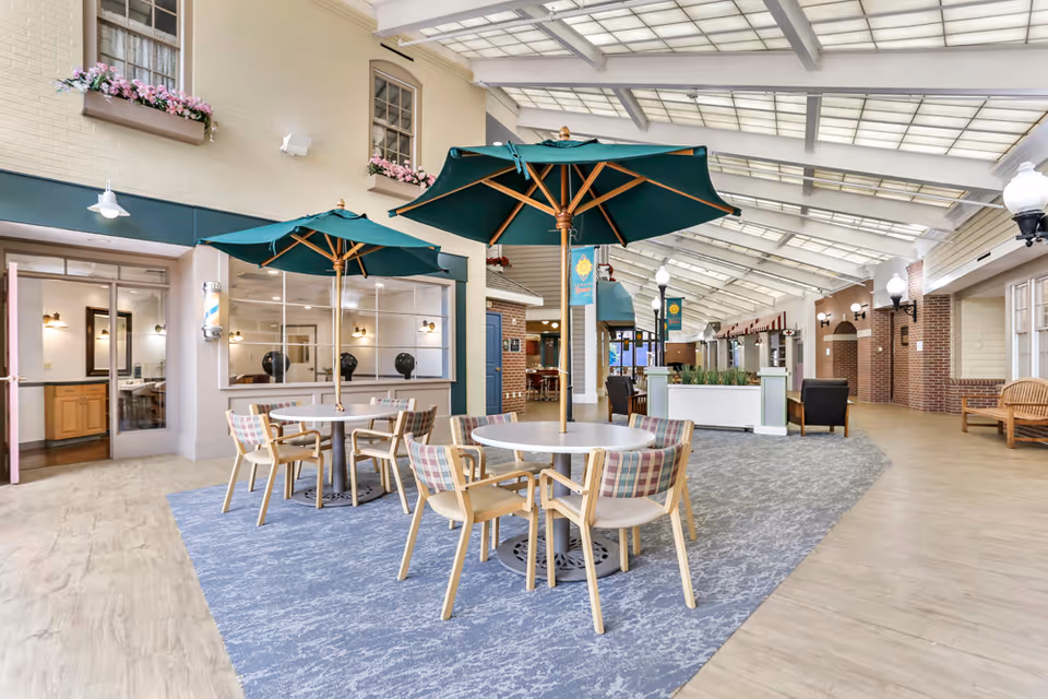Indoor common area with round tables and chairs under green umbrellas, a blue patterned carpet, and a high ceiling with skylights. The space has a cozy, inviting atmosphere with brick and light-colored walls, decorative flower boxes, and various seating options including armchairs and benches.